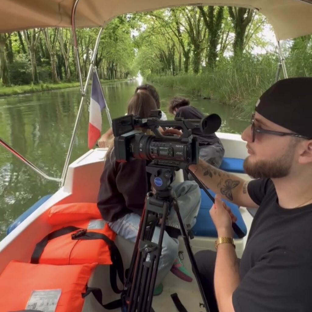 Un vidéaste et photographe à bordeaux et son équipe sur un bateau filmant avec une caméra Sony 4K sur un canal bordé d'arbres.