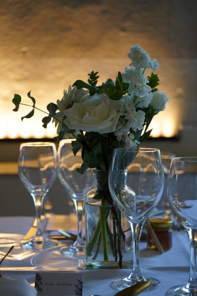 Centre de table composé d'un bouquet de roses blanches et d'eucalyptus dans un vase en verre, entouré de verres à pied sur une nappe blanche avec un éclairage chaleureux en arrière-plan, immortalisé par un Photographe professionnel à Bordeaux.