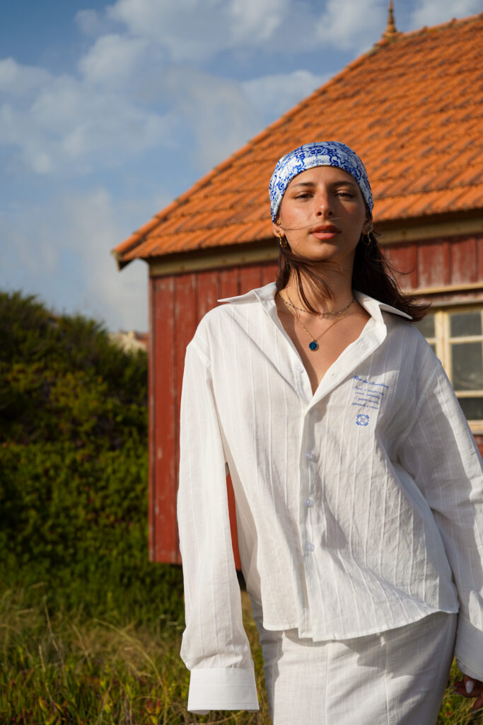 Portrait d'une jeune femme portant un bandana bleu et blanc et une chemise blanche texturée, posant devant une cabane en bois rouge sous un ciel ensoleillé, capturé par un Photographe professionnel à Bordeaux.