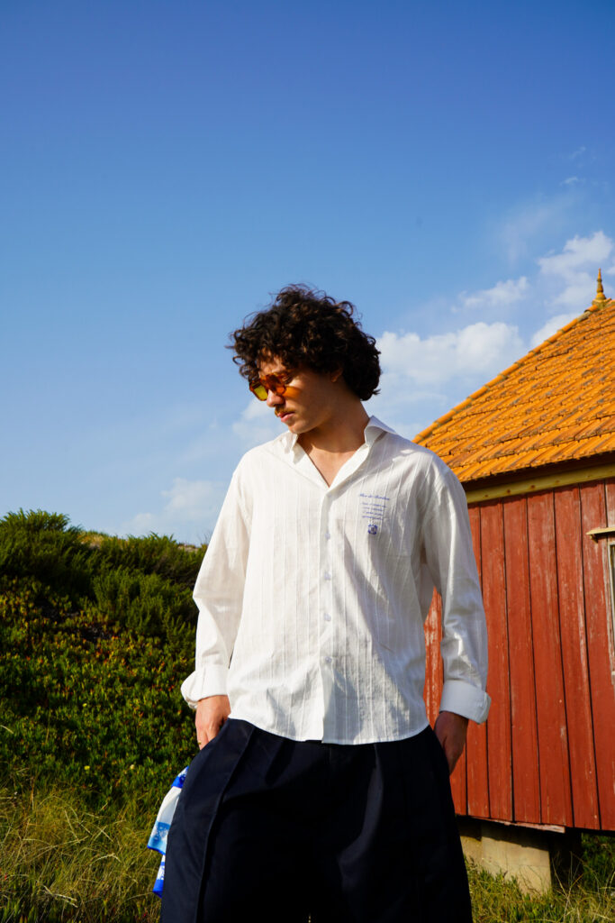 Portrait d'un homme aux cheveux bouclés portant une chemise blanche texturée et des lunettes teintées orange, posant devant une cabane en bois rouge sous un ciel bleu, réalisé par un Photographe professionnel à Bordeaux.